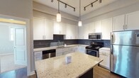 Located at 10504 Castile Rd in Henrico, Virginia, this single-floor residence offers a thoughtfully designed layout with modern finishes throughout. The kitchen is a standout feature, showcasing crisp white shaker cabinetry, a striking gray subway tile backsplash, and light granite countertops with speckled detailing. A dark-cabinetry island with matching granite provides additional prep and dining space, illuminated by stylish pendant lighting. Stainless steel appliances, including a gas range, built-in microwave, dishwasher, and refrigerator, complete the well-appointed kitchen. Track lighting adds a contemporary touch overhead. The open layout connects the kitchen seamlessly to the living area, which features rich hardwood-style flooring, a tray ceiling with a ceiling fan, and glass-paned French doors that invite natural light. A distinctive sliding barn door adds a modern design element. The bedrooms are carpeted for comfort, each with tray ceilings, ceiling fans, and large windows offering views of surrounding greenery. The bathrooms are clean and functional, featuring tub/shower combos with chrome fixtures and dark vanity cabinetry. A dedicated laundry room with side-by-side washer and dryer and overhead dark wood cabinetry adds everyday convenience to this well-organized residence.