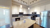 Located at 10504 Castile Rd in Henrico, Virginia, this single-floor residence offers a thoughtfully designed layout with modern finishes throughout. The kitchen is a standout feature, showcasing crisp white shaker cabinetry, a striking gray subway tile backsplash, and light granite countertops with speckled detailing. A dark-cabinetry island with matching granite provides additional prep and dining space, illuminated by stylish pendant lighting. Stainless steel appliances, including a gas range, built-in microwave, dishwasher, and refrigerator, complete the well-appointed kitchen. Track lighting adds a contemporary touch overhead. The open layout connects the kitchen seamlessly to the living area, which features rich hardwood-style flooring, a tray ceiling with a ceiling fan, and glass-paned French doors that invite natural light. A distinctive sliding barn door adds a modern design element. The bedrooms are carpeted for comfort, each with tray ceilings, ceiling fans, and large windows offering views of surrounding greenery. The bathrooms are clean and functional, featuring tub/shower combos with chrome fixtures and dark vanity cabinetry. A dedicated laundry room with side-by-side washer and dryer and overhead dark wood cabinetry adds everyday convenience to this well-organized residence.