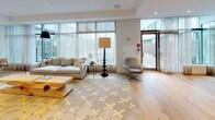 This thoughtfully designed library space combines contemporary architecture with warm, curated details across a single open floor. Light oak hardwood flooring runs throughout, anchoring the space with a clean, natural foundation. A striking linear fireplace is set flush against a crisp white wall, serving as a dramatic focal point that divides the lounge and dining areas. Floor-to-ceiling windows and glass doors line the perimeter, inviting abundant natural light and offering views of the surrounding outdoor environment, while sheer curtains provide a soft, refined touch.

The lounge area features a comfortable neutral sofa, an eclectic plaid swivel chair, and a sculptural wood coffee table atop a bold geometric patterned rug. A glass-enclosed meeting room, labeled "MEET," is outfitted with a long conference table, pendant lighting, and a wall-mounted display screen, offering a semi-private collaborative zone within the open layout.

Built-in rustic wood shelves, finished with a live-edge aesthetic, showcase an organized collection of books, framed photos, decorative objects, and a charming model airplane, adding personality and warmth. Recessed lighting and carefully selected furnishings complete the polished, welcoming ambiance.