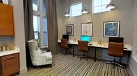 This single-floor space is designed as a functional yet stylish work area, offering a well-appointed environment suited for focused productivity. The room features soaring ceilings that amplify the sense of openness, complemented by tall, floor-to-ceiling windows along one wall that flood the space with natural light. Additional clerestory windows near the ceiling further enhance brightness throughout the day.

A long, streamlined white desk runs along one wall, accommodating two workstations each equipped with a computer monitor and brown leather rolling chairs. Industrial-style chrome pendant lights hang overhead, providing targeted task lighting. Gold-toned desk accessories and articulating brass lamps add decorative accents to the workspace.

Framed blueprint-style artwork depicting vintage inventions adorns the gray walls, contributing an intellectual and creative atmosphere. A patterned wingback chair positioned near the windows provides a comfortable seating alternative for reading or informal conversation. The space is grounded by a striped, multi-toned area rug that ties together the neutral color palette. Overall, the room balances a professional, modern aesthetic with warm, inviting details.