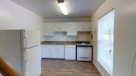 This 2-floor space offers a practical and straightforward layout designed for everyday living. The kitchen is a notable feature, outfitted with white cabinetry that lines the walls, providing ample storage above and below the dark-toned countertops. A stainless sink sits centrally along the counter, flanked by a white dishwasher and a gas range, while a full-size refrigerator anchors the far end of the kitchen. The wood-style flooring adds warmth and visual continuity throughout the space. A large window with horizontal blinds allows generous natural light to fill the kitchen, creating a bright and airy atmosphere. Overhead, a flush fluorescent light fixture provides consistent, even illumination. The walls are finished in a clean, neutral white, keeping the space feeling open and easy to personalize. The overall design is functional and uncluttered, with a focus on utility and practicality. Spanning two floors, the property offers additional living space beyond the kitchen, making it a well-rounded option for those seeking a straightforward, move-in-ready environment.