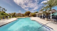Located at 17800 Murdock Cir in Port Charlotte, Florida, this single-level community amenity space is centered around a generously sized outdoor swimming pool that serves as the focal point of the entire area. The pool features a classic geometric design with clearly marked entry steps and is complemented by a separate octagonal spa, providing options for both active swimming and leisurely soaking.

Surrounding the pool is a broad, open concrete sun deck furnished with lounge chairs and shaded umbrella seating, offering ample space for sunbathing and outdoor relaxation. The deck connects seamlessly to a covered patio area adjacent to the main clubhouse building, which features a charming architectural design with a cupola roofline, colonial-style windows, and French doors that open to the outdoor space. Blue lounge seating on the covered patio adds a welcoming touch for shade-seekers.

The outdoor area also includes a dedicated grilling station and picnic space, as well as a children's playground visible in the background, making it a well-rounded recreational destination for all ages. Black wrought-iron fencing encloses the entire pool and amenity area, ensuring security while maintaining open sightlines to the surrounding mature trees and green landscaping. The abundant Florida sunshine and lush natural backdrop create a bright, inviting atmosphere throughout.