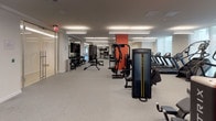 This expansive fitness center is thoughtfully organized into distinct workout zones, each designed to support a comprehensive range of training modalities. The facility is characterized by a clean, modern aesthetic, featuring a neutral white and grey palette accented by bold orange-red column details that provide visual anchoring throughout the open floor plan.

A dedicated indoor cycling studio occupies one end of the facility, finished with warm hardwood flooring and rich wood-paneled walls. Rows of red stationary bikes face mounted flat-screen displays, creating an immersive group cycling environment. Full-length mirrored walls amplify the sense of space and allow for form monitoring.

The main strength training area transitions seamlessly from the cycling studio and features grey carpet flooring throughout. This zone is comprehensively equipped with Matrix cable machines, multi-functional weight racks, adjustable benches, and a full dumbbell rack. A functional training zone is also present, stocked with TRX suspension systems, medicine balls, kettlebells, and balance tools organized on dedicated storage racks.

Treadmills and additional cardio equipment line one side of the main floor, while bold cycling-themed wall graphics add an energetic visual element. Overhead linear fluorescent lighting ensures the entire space is uniformly and brightly illuminated, contributing to a professional, high-performance atmosphere.