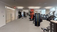 This expansive fitness center is thoughtfully organized into distinct workout zones, each designed to support a comprehensive range of training modalities. The facility is characterized by a clean, modern aesthetic, featuring a neutral white and grey palette accented by bold orange-red column details that provide visual anchoring throughout the open floor plan.

A dedicated indoor cycling studio occupies one end of the facility, finished with warm hardwood flooring and rich wood-paneled walls. Rows of red stationary bikes face mounted flat-screen displays, creating an immersive group cycling environment. Full-length mirrored walls amplify the sense of space and allow for form monitoring.

The main strength training area transitions seamlessly from the cycling studio and features grey carpet flooring throughout. This zone is comprehensively equipped with Matrix cable machines, multi-functional weight racks, adjustable benches, and a full dumbbell rack. A functional training zone is also present, stocked with TRX suspension systems, medicine balls, kettlebells, and balance tools organized on dedicated storage racks.

Treadmills and additional cardio equipment line one side of the main floor, while bold cycling-themed wall graphics add an energetic visual element. Overhead linear fluorescent lighting ensures the entire space is uniformly and brightly illuminated, contributing to a professional, high-performance atmosphere.