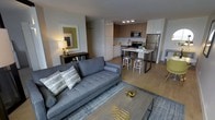 This contemporary single-floor residence offers a thoughtfully designed open-concept layout that seamlessly connects the living and kitchen areas. The living space is anchored by a tufted gray sofa, complemented by a sleek coffee table with gold decorative accents and a textured area rug. A warm table lamp and a bold geometric wall artwork in olive and earth tones add character and color to the space.

The kitchen is outfitted with modern two-tone cabinetry — combining warm wood-finish uppers with clean white lower units — and is equipped with stainless steel appliances, including a refrigerator, range, and built-in microwave. A peninsula-style breakfast bar with a white countertop and industrial-style bar stools serves as both a functional prep area and a casual dining spot, creating a natural transition between the kitchen and living zone.

Light wood-style flooring runs throughout, contributing to a bright, cohesive aesthetic. The overall ambiance is clean and contemporary, with a carefully curated palette of grays, whites, warm wood tones, and gold accents that give the space a polished yet approachable feel. The open layout maximizes the sense of space, making the interior feel airy and well-proportioned.