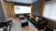 This single-floor space makes a strong visual impression through its carefully curated charcoal and gold color palette. The living area serves as the focal point of the space, anchored by a sleek black leather sofa accented with mustard yellow and grey throw pillows, complemented by a matching black leather ottoman that doubles as additional seating or a footrest. A patterned accent chair adds a touch of visual contrast and texture to the seating arrangement.

Floor-to-ceiling gold velvet curtains frame the large, multi-pane windows that span the width of the room, allowing generous amounts of natural light to filter in while reinforcing the warm tonal theme. The deep charcoal grey walls create a bold, sophisticated backdrop that ties the furnishings together cohesively.

Underfoot, light neutral carpeting softens the overall aesthetic and adds comfort throughout. A small side table positioned near the seating area provides functional surface space. The layout is thoughtfully arranged to maximize both comfort and flow, creating an intimate yet open atmosphere. The interplay of dark walls, warm gold accents, and abundant natural light gives this space a modern, polished ambiance suited for relaxed living.