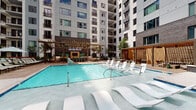 Located at 417 W Peace St in Raleigh, NC, this thoughtfully designed outdoor amenity level offers a well-rounded collection of communal spaces suited for relaxation and recreation. The centerpiece is a resort-style swimming pool with in-water lounge chairs and a spacious sun deck, flanked by a pergola-covered outdoor bar and entertainment area equipped with a mounted television, creating an ideal setting for both casual gatherings and leisure.

The courtyard extends beyond the pool to reveal multiple distinct zones, all unified by a refined mix of brick, wood, and glass architecture characteristic of modern urban construction. Paver walkways guide visitors through lush planted beds and sculptural greenery, connecting covered lounge areas furnished with stylish woven pod chairs and generous sectional sofas. A ping pong table adds a playful recreational element to the open-air terrace.

One standout feature is a warm, wood-paneled wall serving as a dramatic backdrop for an outdoor living room complete with a wall-mounted TV and cushioned seating — ideal for outdoor entertaining. Colorful sculptural wall art accents the brick facade, lending an artistic character to the space. The multi-story residential building rises on all sides, creating an enclosed yet open-sky courtyard atmosphere that balances privacy with a vibrant community feel.