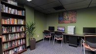 This single-floor business center offers a functional and well-appointed workspace designed to meet everyday computing and printing needs. The room features sage green walls that contribute a calm, professional atmosphere, complemented by a dark drop ceiling with recessed panel tiles that lend a polished, office-like feel underfoot, patterned carpet in muted tones adds texture and comfort to the space.

Along one wall, a continuous countertop workstation accommodates two desktop computer monitors positioned side by side, with ergonomic, contemporary chairs featuring curved backs and warm wood-toned accents providing comfortable seating. A laser printer sits at the end of the counter, making the station a fully equipped work area. A tall, slim bookshelf along the adjacent wall holds an organized collection of books and resources, adding both utility and visual interest.

A large, colorful mosaic-style artwork hangs prominently above the workstations, serving as a focal point that introduces warmth and creativity into the room. A tall potted tropical plant in a dark square planter enhances the space with a touch of nature. Overall, the business center balances practicality with inviting design elements.