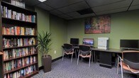 This single-floor business center offers a functional and well-appointed workspace designed to meet everyday computing and printing needs. The room features sage green walls that contribute a calm, professional atmosphere, complemented by a dark drop ceiling with recessed panel tiles that lend a polished, office-like feel underfoot, patterned carpet in muted tones adds texture and comfort to the space.

Along one wall, a continuous countertop workstation accommodates two desktop computer monitors positioned side by side, with ergonomic, contemporary chairs featuring curved backs and warm wood-toned accents providing comfortable seating. A laser printer sits at the end of the counter, making the station a fully equipped work area. A tall, slim bookshelf along the adjacent wall holds an organized collection of books and resources, adding both utility and visual interest.

A large, colorful mosaic-style artwork hangs prominently above the workstations, serving as a focal point that introduces warmth and creativity into the room. A tall potted tropical plant in a dark square planter enhances the space with a touch of nature. Overall, the business center balances practicality with inviting design elements.
