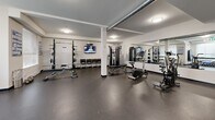 Located at 691 Hoylake Ave in Langford, BC, this fitness centre offers a generously proportioned, single-floor workout space designed to support a full range of exercise routines. The room features a clean, modern aesthetic with crisp white walls, structural columns, and durable rubber flooring suited to high-traffic athletic use. Exposed ventilation ductwork runs along the ceiling, adding an industrial character to the space while ceiling-mounted flush lights provide consistent, even illumination throughout.

The cardio zone is anchored by a row of Matrix treadmills and additional cardio machines, each equipped with individual monitors for entertainment during workouts. The strength training area includes a cable machine, squat rack with weight plates, adjustable benches, free weights, and a stationary bike, offering a comprehensive selection for varied training needs. Wall-to-wall mirrors along one side of the room visually expand the space and assist users in monitoring their form.

Natural light filters in through windows on one end of the room, with a glass-panelled exit door providing views of greenery beyond. Seating is available along the walls, and informational boards are posted for user guidance. The overall ambiance is functional, bright, and well-organized, making this fitness centre a practical and inviting facility for residents in the Langford community.