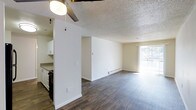 This single-floor residence offers a clean, functional layout that maximizes the sense of openness throughout. The main living area is defined by rich, dark wood-look flooring that extends seamlessly across the space, creating a warm and cohesive feel underfoot. A textured popcorn ceiling adds dimension overhead, while a ceiling fan with a globe light fixture provides both airflow and ambient lighting to the central area.

The kitchen is partially open to the living space, separated by a short dividing wall that maintains a sense of connectivity. White cabinetry lines the kitchen walls, offering ample storage, while dark appliances provide a clean contrast. A baseboard heater runs along the far wall of the living area, ensuring efficient climate control throughout the colder months.

Natural light is a standout feature of this space, with a large sliding glass door positioned at the end of the living area, drawing in daylight and offering a view of the surrounding greenery. The neutral white walls throughout provide a versatile backdrop, making the space easy to personalize. The overall ambiance is bright, clean, and move-in ready, offering a comfortable and practical living environment.