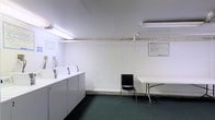 This single-floor laundry facility is designed with practicality and efficiency in mind. The space features a row of top-loading washing machines lined along one wall, providing ample capacity for residents or users. The painted white cinder block walls give the room a clean, utilitarian appearance, while the dark green carpet flooring adds a degree of comfort underfoot. Overhead fluorescent lighting and track lighting ensure the entire space is brightly and evenly illuminated. A folding table and chair are positioned near the center of the room, offering a convenient surface for sorting and folding laundry. Informational notices are posted on the walls, keeping users informed of facility rules and guidelines. The layout is open and uncluttered, allowing for easy movement throughout the space. Overall, the facility presents a straightforward, no-frills environment that prioritizes functionality, cleanliness, and ease of use.
