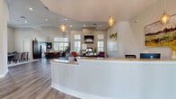 This well-appointed community clubhouse offers a thoughtfully designed single-floor layout spanning a generous footprint. Upon entry, visitors are greeted by a curved granite-topped reception desk accented by elegant pendant lighting and warm hardwood-style flooring that flows throughout the space. Soaring ceilings and an abundance of natural light from multiple windows create an open, airy ambiance.

The combined kitchen and living room area serves as the heart of the clubhouse. The kitchen features dark espresso cabinetry, granite countertops, stainless steel appliances, and a Starbucks coffee station — ideal for resident gatherings. The adjacent living area centers around a dramatic stone fireplace with a mounted flat-screen television, flanked by large windows that flood the room with natural light.

The exercise room is well-equipped with treadmills, ellipticals, stationary bikes, and a free-weight station. Floor-to-ceiling windows overlook the outdoors, while motivational typographic murals energize the space.

A hallway connects the interior rooms to additional amenities. Outside, a covered patio lounge features a matching stone fireplace, comfortable seating, and a gated entry leading directly to a resort-style swimming pool surrounded by lounge chairs and lush landscaping.
