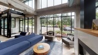 Located at 1367 Willow Street in Minneapolis, Minnesota, this contemporary lobby space makes a striking first impression through its bold industrial-modern design. Exposed concrete columns and beams rise dramatically overhead, framing expansive floor-to-ceiling windows that flood the interior with abundant natural light and offer open views of the surrounding greenery and streetscape.

The main lounge area anchors the space with a generous sectional sofa in deep navy blue, complemented by tufted gray accent chairs and a central fireplace, creating a welcoming and sophisticated gathering environment. Light wood-plank flooring runs throughout, softening the industrial concrete aesthetic and adding warmth.

A secondary seating area features tall, curved wingback chairs in slate blue with vibrant yellow accent pillows, arranged around natural wood side tables. Warm-toned wood cladding on structural columns and partition walls introduces an organic richness that contrasts beautifully with the building's raw concrete framework.

The entrance is highlighted by a bold, large-scale abstract mural in teal, yellow, and blush tones, positioned beneath a sleek open-riser staircase with glass railings — a true focal point that adds artistic character to the space. Together, these elements create a polished, inviting environment that balances urban industrial architecture with comfortable, thoughtfully curated design.