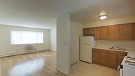 This single-floor space offers a practical and comfortable layout designed for easy everyday living. The kitchen is positioned at the entry point of the unit and is equipped with warm wood-toned cabinetry, light tile flooring, generous counter space, and a full-size refrigerator. The kitchen flows naturally into the adjoining living area, which is separated by a partial dividing wall that adds subtle definition without closing off the space. The living area features light hardwood floors that lend warmth and continuity throughout, while a large window along the far wall allows natural light to pour in, brightening the entire space. Baseboard heating runs along the perimeter walls, ensuring year-round comfort. The neutral, cream-toned walls throughout the unit provide a clean and versatile backdrop, making the space easy to personalize. The overall ambiance is bright, functional, and welcoming — a solid single-floor layout that balances open-concept living with clearly defined zones for cooking and relaxing.