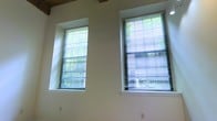 This single-floor space presents a clean, open layout characterized by smooth, light-colored walls and notably high ceilings that contribute to an airy, spacious atmosphere. The ceiling structure, partially visible in the upper corners, hints at an industrial or loft-inspired architectural character, adding a distinctive design element to the overall aesthetic.

Natural light is a defining feature of this space. Two tall, generously sized windows are fitted with horizontal venetian blinds and frame views of lush, leafy greenery outside, bringing a sense of the outdoors into the interior. The windows are set into the walls with a slight recess, adding depth and architectural interest to the wall plane. The varying heights of the windows — one set higher and one at a more standard level — reflect a thoughtful approach to both design and light distribution throughout the space.

The neutral wall palette and clean finishes create a versatile, move-in-ready environment suitable for a variety of uses. The combination of high ceilings, abundant natural light, and outdoor views makes this a bright, welcoming space with a calm and functional ambiance.