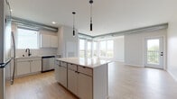 This 2-bedroom, 2-bathroom unit at Beyond Woodbury delivers a thoughtfully designed, single-floor layout with a clean, contemporary aesthetic throughout. The open-concept living and kitchen area is a standout feature, anchored by a large central island with a light quartz countertop and sleek pendant lighting overhead. Exposed ductwork adds an industrial touch to the space, while light hardwood-style flooring flows seamlessly through the main living areas. The kitchen is well-appointed with white cabinetry, a herringbone tile backsplash, stainless steel appliances including a refrigerator, microwave, gas range, and dishwasher, complemented by a matte black faucet and hardware. Floor-to-ceiling windows and a glass balcony door in the living area flood the space with natural light and offer open outdoor views. Both bedrooms are carpeted and generously sized, each featuring large windows that continue the theme of natural light and scenic sightlines. The two bathrooms are finished in a consistent modern style, featuring gray vanities with white countertops, matte black fixtures, large mirrors, and light tile flooring. One bathroom includes a shower-tub combo. This unit combines modern finishes with a bright, airy feel throughout.