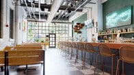 This single-floor dining establishment showcases a thoughtfully designed interior that blends industrial and contemporary aesthetics. The space is anchored by a long bar running along one side, lined with woven rattan-style barstools and backed by sage-green tiled walls with decorative shelving displaying bottles and greenery. A mounted TV and point-of-sale terminals complete the functional bar setup.

The main dining area features a generous run of wooden bench-style booth seating paired with light wood tables, offering a casual yet inviting atmosphere. A separate, more intimate seating section toward the rear includes woven chairs set against whitewashed brick walls adorned with a decorative shelf displaying an array of dried floral arrangements, adding a warm, artisanal touch.

Underfoot, a bold checkerboard tile floor in muted grey and white tones runs throughout the space, lending a retro character that complements the exposed overhead ductwork and open-ceiling industrial infrastructure above. Large steel-framed windows and a glass-paneled entry facade at the front flood the interior with natural light and provide views of surrounding greenery. Lush potted plants near the entrance further enliven the space. Track lighting and pendant bulbs provide warm, layered illumination throughout, creating an energetic yet comfortable dining environment.