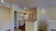 This single-floor residence offers a thoughtfully arranged layout designed for comfortable everyday living. The kitchen serves as a central highlight, featuring light wood cabinetry, a white electric range, built-in microwave, and a full-size refrigerator. A decorative wine rack built into the upper cabinetry adds a charming touch. Track lighting illuminates the workspace efficiently, while recessed lighting enhances the overall brightness of the area. A peninsula breakfast bar with a dark laminate countertop extends into the adjoining living space, creating a natural division between the kitchen and the main living area while maintaining an open, connected feel. Dark hardwood-style flooring runs through the kitchen, complementing the warm neutral tones of the walls. A spacious walk-in pantry or closet is conveniently located just off the kitchen, outfitted with wire shelving for ample storage. Louvered bifold doors are visible in the background, suggesting additional storage or utility areas. The transition from hard flooring to carpet marks the entry into the living space, adding warmth and comfort underfoot. Overall, the space presents a practical and inviting environment with a cohesive, neutral color palette throughout.
