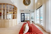 Located in Salem, MA, the Lore lobby is a thoughtfully designed, single-floor space that makes a striking first impression. Soaring double-height ceilings amplify the sense of openness, while floor-to-ceiling windows along the exterior walls flood the interior with abundant natural light and offer views of the surrounding cityscape. Light-toned hardwood flooring runs throughout the main entry area, complementing the bright white walls and creating a clean, modern aesthetic. A distinctive curved, latticed wood partition serves as a defining architectural feature, elegantly separating the entry zone from the interior lounge without sacrificing visual connectivity. A large circular branded medallion anchors the wall above, adding a bold decorative focal point. The welcoming lounge area centers around a richly upholstered terracotta-red sofa, paired with accent chairs and small side tables that introduce warmth and character. Black-framed glass entry doors provide a sleek contrast to the warm interior palette. Beyond the entryway, a well-appointed lounge and amenity area features a curved light wood reception desk with a dark stone countertop, plush rust-toned seating, a bar counter with marble backsplash and wood barstools, and layered lighting including pendant fixtures and recessed ceiling lights. The overall ambiance balances contemporary sophistication with inviting warmth.