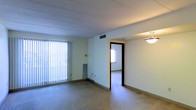 This single-story space features a straightforward, functional layout with neutral-toned walls and wall-to-wall carpeting throughout. The main living area is open and modestly sized, benefiting from a large window dressed with vertical blinds that allow natural light to filter in, brightening the interior. A ceiling-mounted light fixture provides additional overhead illumination. HVAC vents are visible in the ceiling, ensuring climate control throughout the space. A doorway with a dark-trimmed frame leads to an adjacent room, which appears to have its own window, suggesting good natural light distribution across the unit. A small baseboard-style heater or radiator unit is positioned along one wall, adding a supplementary heat source. The ceiling features a subtle textured finish with linear detailing, adding a minor architectural element to the otherwise simple design. The overall ambiance is clean and utilitarian, with a neutral color palette of white and beige tones that create a blank canvas well-suited for personalization. The single-floor layout offers convenient, step-free access throughout the entire space.
