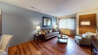 This single-floor residence offers a thoughtfully designed layout with a cohesive aesthetic defined by warm hardwood flooring, soft blue-gray walls, and natural wood accents throughout. The living room serves as the heart of the home, featuring rich hardwood floors, a two-tone color palette of slate blue and warm cream, and generous natural light filtered through blinds and decorative curtains. The space comfortably accommodates a full seating arrangement alongside a media area with a dedicated accent chair and floor lamp.

The dining area is positioned adjacent to the living room and includes a pendant light fixture overhead, sliding wood-panel closet doors, and ample space for a round dining table and multiple chairs. The galley-style kitchen is well-equipped with stainless steel appliances — including a refrigerator, electric range, microwave, and dishwasher — complemented by warm maple cabinetry, light countertops, and hardwood flooring.

The bedroom is a serene retreat with plush carpet, a ceiling fan, a sliding wood closet, natural light from a window with curtains, and coordinated dark wood furniture. The full bathroom features a tub-shower combination with a decorative curtain, a wood-vanity with a single sink, hardwood flooring, and safety grab bars near the tub, adding a practical touch to the design.