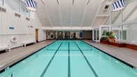 Located at 17 Captains Landing in Strawberry, CA, this single-story indoor aquatic facility is defined by its striking architectural design and refined coastal character. The centerpiece is a large, multi-lane lap pool with vivid aquamarine water and clearly marked lane lines, set beneath dramatically vaulted, white-painted beam ceilings that rise to impressive heights and flood the space with a sense of openness and volume. Clerestory windows at the peak of the gabled roofline allow natural light to pour in, enhancing the bright and airy atmosphere throughout.

The pool deck is finished in neutral tile and is lined with benches along the walls, providing practical poolside seating. At one end of the facility, a generous lounge area features a built-in, L-shaped wood bench with cushioned seating, flanked by stepped wooden planters filled with lush tropical greenery. Floor-to-ceiling glass walls in this area frame panoramic views of the surrounding marina, seamlessly connecting the interior to the waterfront setting outside. Additional amenities visible throughout the space include safety equipment, wall-mounted lighting fixtures in a nautical blue accent, and access to adjacent rooms. The overall ambiance is clean, functional, and elegantly appointed.