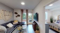 Located at 1601 Broadway in San Diego, unit #324 is a single-floor residence that blends contemporary design with bohemian character. The open-concept layout connects the living area and kitchen seamlessly, with warm hardwood-style flooring running throughout the main spaces.

The living room is bright and inviting, featuring a sliding glass door that opens to a private balcony overlooking lush palm trees. Tall draping curtains frame the greenery views, while decorative wall fans and macramé accents add an eclectic, artful touch. A wall-mounted flat-screen TV and a glass-top coffee table complete the comfortable, stylish space.

The kitchen is well-appointed with light flat-front cabinetry, white quartz countertops, stainless steel appliances including a gas range, refrigerator, and dishwasher, and a peninsula with bar seating illuminated by pendant lighting. Ample cabinetry provides generous storage.

The bedroom features a distinctive whitewashed brick accent wall paired with a rattan headboard, creating a warm, textural focal point. A second sleeping area benefits from a large window with palm tree views and neutral carpeted flooring.

The bathroom offers a clean, modern aesthetic with light wood cabinetry, a white quartz vanity, a framed mirror, and dark tile flooring, all contributing to a polished, cohesive finish throughout the residence.