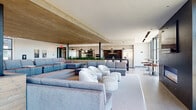 This expansive single-floor amenity space offers a sophisticated, open-concept layout designed for both social gatherings and relaxed lounging. The centerpiece of the living area is a dramatic wood-paneled ceiling that extends over a sunken lounge zone, creating an intimate atmosphere within the larger open floor plan. Generously scaled sectional sofas in soft grey tones are complemented by sculptural white accent chairs and rounded ottomans, offering a variety of comfortable seating arrangements.

A full kitchen anchors one end of the space, equipped with a large island featuring dark granite countertops, bar seating, stainless steel appliances, and sleek pendant lighting. Adjacent to the kitchen, a row of bar-height seating runs alongside the perimeter, ideal for casual dining or socializing.

Floor-to-ceiling glass curtain walls flood the space with abundant natural light and frame panoramic urban views. A bold dark accent wall with a built-in linear fireplace and wall-mounted television adds a striking focal point in the lounge area. A separate dining zone features a substantial wood farmhouse table, flanked by a richly textured horizontal wood plank wall. Light wood-toned flooring and crisp white walls unify the space, lending it a clean, contemporary aesthetic throughout.