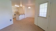 This single-floor unit presents an open-concept layout that seamlessly connects the living area and kitchen. The living space is finished with neutral-toned carpeting, creating a warm and understated ambiance throughout. A front entry door with a sidelight window allows natural light to filter into the space, brightening the interior. The kitchen is well-equipped with white cabinetry, a white refrigerator, and a white range, all arranged efficiently along the back wall. A peninsula counter with an integrated sink and bar-style seating separates the kitchen from the living area, offering both additional prep space and casual dining options. A pendant light fixture hangs above the peninsula, providing focused task lighting, while a fluorescent fixture overhead ensures the kitchen remains well-lit. The transition between the carpeted living area and the kitchen's hard flooring is clean and defined. The overall design is straightforward and functional, with neutral wall tones and simple finishes contributing to a bright, easy-to-personalize living environment suitable for a variety of lifestyles.