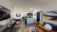 This well-appointed single-floor unit offers a contemporary open-concept layout that seamlessly connects the living, dining, and kitchen areas. The living space features wood-look flooring throughout, a large flat-screen TV setup, and a front entry door flanked by a natural-light-admitting window. A stylish pendant light fixture and circular mirror accent the dining area, which flows directly into the kitchen. The kitchen is outfitted with dark cabinetry, stainless steel appliances including a refrigerator, microwave, and electric range, plus a central island with a white countertop ideal for meal prep and casual dining. The unit includes multiple bedrooms, each with wood-look flooring, ceiling fans, and closets. One bedroom features a dedicated walk-in closet and direct access to a private en-suite bathroom with a vanity, tub-shower combination, and tiled flooring. Additional bedrooms offer natural light through windows with blinds and curtains, alongside generous storage. Two full bathrooms serve the unit, both featuring dark-framed cabinetry, white countertops, tub-shower combinations, and large-format tile flooring. Bold, modern interior doors with satin hardware appear throughout, reinforcing the unit's sleek, urban aesthetic.