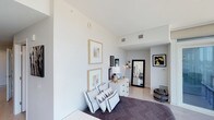 This single-floor bedroom showcases a modern aesthetic through its crisp white walls and light hardwood-style flooring, creating a bright and airy atmosphere. The room benefits from notably tall ceilings, which enhance the sense of spaciousness and openness throughout. A carefully curated gallery wall displays two gold-framed botanical prints, complementing the room's warm neutral tones. A white dresser with gold hardware serves as a functional focal point, topped with a classic white table lamp and decorative accents. A large full-length mirror in a bold black frame leans against the wall, adding depth and dimension to the space. The bed is dressed in a dark patterned duvet, layered with an assortment of geometric-print throw pillows in monochrome tones, adding visual interest. A brass arc floor lamp provides ambient lighting, while a striking black and gold abstract artwork decorates the far wall. Woven basket accessories introduce natural texture. The room's overall design balances contemporary minimalism with carefully chosen decorative elements, resulting in a cohesive, sophisticated living space.