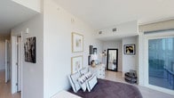 This single-floor bedroom showcases a modern aesthetic through its crisp white walls and light hardwood-style flooring, creating a bright and airy atmosphere. The room benefits from notably tall ceilings, which enhance the sense of spaciousness and openness throughout. A carefully curated gallery wall displays two gold-framed botanical prints, complementing the room's warm neutral tones. A white dresser with gold hardware serves as a functional focal point, topped with a classic white table lamp and decorative accents. A large full-length mirror in a bold black frame leans against the wall, adding depth and dimension to the space. The bed is dressed in a dark patterned duvet, layered with an assortment of geometric-print throw pillows in monochrome tones, adding visual interest. A brass arc floor lamp provides ambient lighting, while a striking black and gold abstract artwork decorates the far wall. Woven basket accessories introduce natural texture. The room's overall design balances contemporary minimalism with carefully chosen decorative elements, resulting in a cohesive, sophisticated living space.