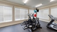 Located at 35 Howard Gap Rd in Fletcher, North Carolina, this fitness center offers a well-equipped, single-floor workout facility designed to support a full range of exercise routines. The space is finished with durable rubber flooring throughout, providing a safe and functional surface for both cardio and strength training activities.

The room is lined with multiple large windows fitted with horizontal blinds, allowing ample natural light to fill the space while maintaining privacy. Crown molding and neutral beige walls lend a clean, polished aesthetic to the interior, complemented by flush fluorescent lighting overhead.

Cardio equipment includes multiple treadmills and elliptical cross-trainers, offering a variety of low- and high-impact workout options. For strength training, the facility is outfitted with several cable and resistance machines featuring red upholstered seats, a multi-function weight stack machine, a leg press unit, and a dumbbell rack with a selection of free weights and a adjustable bench. Exercise balls are also available for core and flexibility work.

Access to the fitness center is provided through a set of glass-paneled French doors, clearly marked with exit signage. The layout is open and organized, allowing users to move comfortably between stations. Overall, the space presents a clean, functional, and well-maintained environment suited for a comprehensive fitness routine.