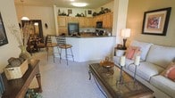 This single-floor residence is designed around an open-concept floor plan that connects the kitchen and living area into one cohesive, flowing space. The kitchen is outfitted with light wood cabinetry extending to the ceiling, providing generous storage capacity, while the upper surfaces above the cabinets are styled with decorative accents. Dark granite-style countertops run along the kitchen perimeter and wrap around a peninsula that doubles as a breakfast bar, accommodating bar-height seating with wrought iron stools. Black appliances complement the cabinetry and countertops. The living area is carpeted in a neutral tone, creating a comfortable and cohesive feel throughout the space. A glass-topped coffee table with ornate metal framing serves as a central focal point, accompanied by seating accented with soft pink toss pillows and a table lamp that adds warm ambient lighting. The open layout allows for easy sightlines between the kitchen and living space, making the overall environment feel connected and well-suited for both everyday living and casual entertaining. The warm wall tones contribute to an inviting, relaxed ambiance throughout.