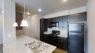 This single-floor residence offers a thoughtfully designed interior with a strong emphasis on modern aesthetics and functional living. The kitchen serves as a standout feature, showcasing rich dark cabinetry that extends to the ceiling, providing generous storage space. Granite-style countertops in a light speckled finish create a striking contrast against the dark lower and upper cabinets, lending the space a polished, contemporary feel. A double stainless steel sink is paired with a matching faucet and accented by a pendant light fixture overhead, adding a refined touch to the workspace. Black appliances — including a range, microwave, and refrigerator — complete the kitchen's cohesive, modern palette. Recessed lighting throughout keeps the space well-lit and visually open. The flooring transitions from wood-style planks in the kitchen area toward the adjoining spaces, contributing warmth and continuity. A view into the adjacent area reveals additional living space and a paneled interior door, hinting at a practical and well-organized layout. The overall ambiance is clean, modern, and move-in ready, with design choices that balance style and everyday functionality seamlessly.