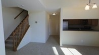 This two-story residential space offers a practical and well-organized layout across its two levels. The main floor features a carpeted living area bathed in natural light, creating a warm and inviting atmosphere. A staircase with classic wooden railings and balusters provides access to the upper level, adding a traditional architectural element to the interior. Adjacent to the living area, a hallway connects the main spaces, illuminated by a flush-mount ceiling light fixture that keeps the corridor bright and welcoming. The kitchen is partially visible from the living area through a recessed opening in the wall, showcasing dark-toned cabinetry and a dark countertop that contrast neatly with the neutral walls. This pass-through design element connects the kitchen to the living space while maintaining a sense of separation between the two areas. Throughout the main floor, the light-colored carpet and clean white walls contribute to an open, airy feel. The overall design is straightforward and functional, emphasizing ease of movement between spaces and a cohesive, neutral color palette that provides a versatile backdrop for a variety of interior styles.