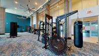 Located at 3110 Grand Ave in Pinellas Park, Florida, this fitness center is a spacious, single-floor facility designed to support a comprehensive workout experience. The space is defined by its soaring double-height ceilings, exposed industrial ductwork, and an abundance of natural light that pours in through floor-to-ceiling windows overlooking lush Florida landscaping. Textured wood-finished columns punctuate the open floor plan, adding a warm, modern-industrial aesthetic to the space.

The flooring throughout features a distinctive mosaic-style carpet in shades of teal, gray, and blue, complementing the bold teal accent walls that energize the environment. A glass-enclosed studio area with hardwood flooring provides a dedicated zone for group or individual training sessions.

The strength training section is outfitted with power racks, weight plates, medicine ball storage, a JumpSport fitness trampoline, and heavy punching bags. The cardio area includes ellipticals, stationary bikes, and stair climbers, while a variety of selectorized cable and resistance machines are distributed throughout the space. Wall-mounted flat-screen TVs and large ceiling fans contribute to member comfort. Full-length mirrors enhance the sense of space and support proper form during workouts.