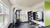 This fitness center offers a thoughtfully organized workout space designed to support a full range of exercise routines. The room is defined by a striking lime-green accent wall that energizes the environment, contrasting with the clean white walls and ceiling throughout the rest of the space. Recessed lighting provides bright, even illumination across the rubber-floored workout area, which is well-suited for high-impact activity.

The cardio section features a treadmill and elliptical trainer, positioned near large windows that allow natural light to filter in and offer views of the surrounding outdoor greenery. A flat-screen television is mounted nearby for entertainment during workouts. Strength training is well-represented with an Inspire functional trainer cable machine, a dedicated weight bench, and a comprehensive rack stocked with dumbbells, medicine balls, and kettlebells in various sizes. A large wall mirror spans a significant portion of one wall, enhancing the sense of openness and allowing users to monitor their form.

The space is accessed via a dedicated exterior door, adding convenience and a sense of privacy. Safety features including an exit sign and fire alarm are present. The overall layout is efficient and purposeful, making effective use of the available footprint to deliver a versatile and motivating fitness environment.