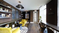 This single-floor space showcases a carefully curated interior that blends industrial and contemporary design elements. The room is anchored by rich, dark hardwood flooring that runs throughout, complemented by deep brown walls that lend a warm, intimate ambiance. Bold yellow upholstered armchairs serve as striking focal points, arranged atop black-and-white geometric rugs that add visual contrast and definition to seating areas.

Overhead, distinctive industrial-style pendant lights with wide, layered metal shades hang from a crisp white ceiling, providing both functional lighting and a design statement. Built-in shelving along the walls offers display space for books and decorative objects, adding character and depth to the room.

Natural light filters in generously through floor-to-ceiling windows dressed with sheer white curtains along one side of the space, creating a bright counterbalance to the darker tones of the interior. A leather rolling chair and a wooden desk occupy the windowed area, suggesting a functional workspace. Lush indoor plants near the windows introduce a natural element, while a glass-paneled door provides a clean architectural transition point within the space.