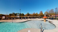 This community amenity space features two distinct areas designed for recreation and wellness. The outdoor pool area boasts a generously sized, freeform swimming pool with clear turquoise water, accessible via metal entry rails. A wide concrete pool deck surrounds the water and is lined with multiple lounge chairs, ideal for relaxing under open skies. The area is enclosed by a black iron fence, with a backdrop of mature trees and expansive green space beyond. A covered gazebo-style structure adds a shaded gathering spot to the deck.

The indoor fitness center is a spacious, well-lit facility featuring high ceilings with crown molding and large windows that invite abundant natural light and views of the surrounding community. The flooring consists of stylish herringbone-patterned carpet throughout. Ceiling fans maintain comfortable airflow across the space. Equipment is comprehensive and includes treadmills with mounted flat-screen televisions, a rowing machine, Precor stationary bike, cable and multi-functional strength machines, a squat rack with bench, a full dumbbell rack, medicine ball storage, battle ropes, resistance bands, stability balls, and a large wall-mounted mirror that amplifies the sense of space. The fitness center reflects a commitment to providing residents with a full range of cardio and strength training options.