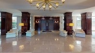 This distinguished lobby makes a memorable first impression with its expansive polished marble tile flooring that reflects the abundant natural light pouring in through dramatic floor-to-ceiling glass walls. The space is thoughtfully divided into distinct zones, seamlessly blending arrival and lounging functions into a cohesive, welcoming environment.

At the heart of the lobby, a striking brass sputnik-style chandelier anchors the central hall, casting a warm glow over the symmetrically arranged seating and rich mahogany architectural elements. Tall, dark-wood column surrounds and paneled cabinet millwork lend a sense of refinement and gravitas, while elevator doors framed in matching mahogany millwork provide an elegant focal point at the rear.

Two comfortable seating lounges flank the main corridor, each thoughtfully appointed with upholstered sofas, accent chairs, and wooden occasional tables. One lounge features floor-to-ceiling wood paneling adorned with a framed nautical painting, exuding a classic, club-like atmosphere. The other opens directly to sweeping views of lush landscaped greenery through expansive glass walls dressed with gold drapery panels, creating an airy, garden-like ambiance.

Potted indoor plants add organic warmth throughout, while the seamless connection to the manicured outdoor surroundings reinforces a sense of tranquility and openness. Overall, the space projects timeless sophistication with a gracious, welcoming character.