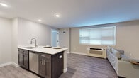 This 2-bedroom, 2-bathroom single-floor residence offers a thoughtfully designed layout that maximizes functionality and comfort. The open-concept kitchen and living area serves as the heart of the home, where a central island with a white countertop and bar seating for two creates a natural gathering space. The kitchen is equipped with stainless steel appliances, including a range, microwave, and refrigerator, complemented by white cabinetry that keeps the space feeling bright and organized. Durable wood-look flooring runs throughout the common areas, adding warmth and continuity.

The living area provides a comfortable retreat with a large window that invites natural light, finished with neutral-toned walls that lend a clean, modern aesthetic. Both bedrooms feature plush carpeting and windows with exterior views, offering a quiet and restful environment. The primary bedroom is a well-proportioned private space, while the second bedroom mirrors its calm ambiance. The two bathrooms are finished in a clean, minimalist style, each featuring a tub-shower combination and wood-look tile flooring. A barn-style sliding door is a notable architectural detail visible in the unit's layout. Recessed lighting throughout ensures consistent, practical illumination across all rooms.