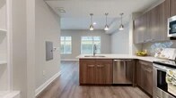 This single-floor unit offers an efficiently organized layout comprising a living room, kitchen, bathroom, and closet. The open-concept kitchen serves as a central feature, equipped with stainless steel appliances including a range, microwave, and dishwasher. Rich walnut-toned cabinetry pairs with white quartz countertops and a geometric tile backsplash, while three brushed nickel pendant lights hang overhead, adding a contemporary touch. A peninsula with an undermount sink connects the kitchen to the living area, promoting an open, airy feel.

The living room benefits from multiple windows fitted with horizontal blinds, allowing natural light to fill the space and offering views of the surrounding exterior. Light-toned wood-style flooring runs consistently throughout the main living areas, contributing to a cohesive, clean aesthetic.

The bathroom is finished with large-format tile flooring, a walnut-toned vanity with white countertop, a full tub-shower combination with white subway tile surround, and a stacked washer and dryer unit conveniently integrated into the space.

The entryway closet features built-in white shelving and coat hooks, providing practical organizational storage upon entry. Neutral gray walls and white trim throughout unify the overall design of the space.