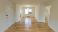 This single-floor space presents a warm and understated aesthetic, anchored by well-maintained hardwood floors that stretch throughout the main living area, reflecting natural light and adding a timeless charm to the interior. The open layout of the living and dining area creates a sense of spaciousness, with cream-toned walls that lend a clean, neutral backdrop adaptable to a variety of interior styles. A classic pendant light fixture hangs above the dining area, providing both functional illumination and a subtle decorative focal point. A window positioned along the far wall allows natural light to filter into the space, complemented by a wall-mounted air conditioning unit for climate control. A radiator beneath the window indicates the presence of a traditional heating system, contributing to year-round comfort. Crisp white baseboards and trim offer a polished contrast to the warm wood flooring throughout. Doorways visible from the main area suggest additional rooms beyond, hinting at a functional and well-proportioned floor plan. Overall, the space exudes a simple, classic character suited for comfortable everyday living.