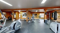 This fitness center combines rustic architectural charm with practical workout functionality. The main exercise area is the heart of the space, distinguished by warm, honey-toned log cabin walls, sturdy exposed wooden support columns, and flush ceiling-mounted fluorescent lighting that ensures the room is well-lit throughout. A large University of Colorado (CU) Buffaloes logo adorns one of the log walls, lending the space a spirited, collegiate character.

The exercise floor is finished with durable dark rubber flooring, ideal for high-traffic fitness use. The room is generously sized to accommodate a wide variety of equipment, including Precor elliptical trainers, a treadmill, a stationary spin bike, a rowing machine, a cable strength machine, a leg press, and additional resistance and free weight stations. Large glass-paned entry doors along one wall allow natural light to filter in and provide views of the surrounding outdoor area.

A full-length mirror is mounted on one wall, enhancing the sense of openness and allowing users to monitor their form. The space also includes an accessible restroom featuring clean white tile wainscoting, wood-look vinyl plank flooring, grab bars, and modern fixtures — ensuring the facility is welcoming and functional for all users. Together, these two areas form a complete, well-appointed fitness facility.