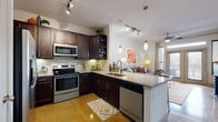 This well-appointed single-floor residence combines modern finishes with a warm, inviting atmosphere throughout. The open-concept kitchen is a standout feature, showcasing rich dark cabinetry, granite countertops, a tile backsplash, and a full suite of stainless steel appliances including a range, microwave, dishwasher, and refrigerator. A central island with a built-in sink and seating provides additional workspace and casual dining. Polished concrete floors extend through the kitchen and living areas, adding an industrial-chic character that contrasts beautifully with the warm tones of the space. Pendant lighting and track lighting illuminate the kitchen, while a ceiling fan keeps the living area comfortable. The living room is bright and open, benefiting from natural light streaming through large French doors that lead to an outdoor area, complemented by transom windows above. The bedrooms offer plush carpeting, generous natural light through wide windows with blinds, and a calm, restful ambiance. The bathroom features a granite-topped dark vanity with an undermount sink and polished concrete flooring, connecting conveniently to the bedroom. Throughout, the residence presents a cohesive design with neutral walls, contemporary fixtures, and thoughtful finishes.