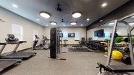 This fitness center is a comprehensively outfitted facility designed to accommodate a wide range of workout styles and fitness goals. The space is organized across a single floor and divided into distinct functional zones, each thoughtfully arranged for both usability and comfort.

The main workout area features a full rack of dumbbells displayed along a floor-to-ceiling mirror wall, maximizing the sense of space while allowing users to monitor their form. A weight bench and cable resistance machine anchor the strength training section, complemented by stability balls and medicine balls for functional fitness. Multiple wall-mounted flat-screen televisions are positioned throughout the space for entertainment during workouts.

A dedicated cardio zone houses a row of treadmills, elliptical trainers, and a rowing machine, bathed in natural light from several windows fitted with blinds. Ceiling fans and recessed lighting provide a well-ventilated and well-lit environment. An additional smaller cardio alcove offers a more private setting with its own elliptical and treadmill, alongside another mounted television.

The flooring throughout is durable rubber-aggregate material, appropriate for heavy equipment and high-traffic use. The color palette blends warm beige walls with bold dark accent walls, lending the space a polished, modern aesthetic. Colorful abstract artwork adds visual interest near the entryway, which is outfitted with a stainless steel waste bin and glass-paneled door.