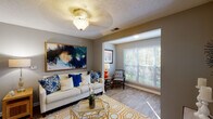 This single-floor residence offers a thoughtfully designed layout that balances comfort and functionality across its key living spaces. The living room serves as the heart of the home, featuring wood-look flooring, a ceiling fan with light fixture, and a sunlit reading nook with large windows that invite natural light and outdoor views. Neutral gray walls provide a versatile backdrop throughout the space.

The kitchen is outfitted with crisp white cabinetry, dark countertops, and stainless steel appliances including a refrigerator, range, and microwave. A peninsula-style counter separates the kitchen from the open dining area, which accommodates a round glass-top table beneath a pendant light fixture. The kitchen also features wood-look flooring that carries through from the main living areas.

Two well-proportioned bedrooms offer carpeted flooring and ceiling fans, each with windows providing natural light. The primary bedroom features a textured ceiling and ample space for a queen or king bed. Two bathrooms serve the residence — one with a tub-shower combination, wood-look flooring, and a vanity with white cabinetry, and a second full bathroom also featuring a tub-shower with decorative accents and wood-look flooring. The overall ambiance is warm, cohesive, and move-in ready.