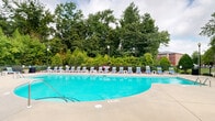 Located at 830 Allonby Rd in Fayetteville, NC, this generously sized community amenity center offers an impressive array of shared facilities across its single primary floor. The exterior is defined by classic red brick construction, with glass-paneled entryways providing views of the expansive outdoor pool area.

The centerpiece of the outdoor space is a large, resort-style swimming pool with a curving edge, surrounded by a broad concrete deck lined with colorful lounge chairs. Mature trees and lush greenery form a natural backdrop, creating a private, park-like setting. A pergola with additional seating adds a shaded gathering spot alongside the pool.

Inside, the amenity building transitions seamlessly from a welcoming reception area featuring warm wood cabinetry, subway tile backsplash, pendant lighting with an industrial flair, and hardwood-style flooring. Crown molding and chair rail detailing throughout lend the interior a polished, well-appointed character. A cozy lounge area with modern blue accent chairs and curated artwork provides a comfortable space for residents to gather.

The fitness center is well-equipped with a full complement of strength and cardio machines, rubber flooring, wall mirrors, and bright overhead lighting. French doors connect it to the main interior corridor for easy access. Completing the amenities is a private screening room outfitted with tiered, theater-style seating and a ceiling-mounted projector, ideal for community movie nights.